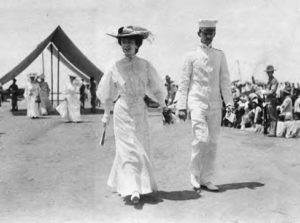 a woman in white dress and fancy hat is walking next to a man in all white.