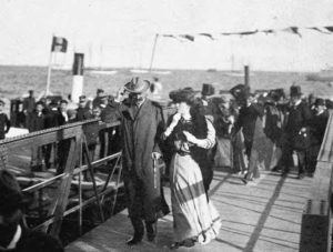 photograph of a man and woman walking on a well-populated dock, with some ships in the water behind them
