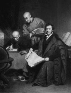 three men sit at a table. One looks over the shoulder of another who is writing. 