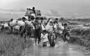 photograph of people walking through mud and standing water while carrying bundles and packs.