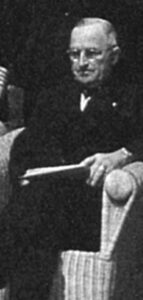 a man in a suit and glasses sits with a stern look on his face