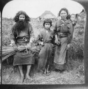 photo of a family consisting of a mother, father, young son, and baby. they wear simple robes and stand in front of long grass and their house in the distance.