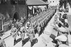 A photograph capturing Nazi German army troops marching through a city, while onlookers along the streets salute them. The image represents a historical scene from the era, depicting the military presence and public response during that time.