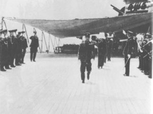 A row of Japanese soldiers standing on either side of a disembarking vessel. Sailors are seen disembarking from the boat, one holds the ashes of Admiral Yamamoto.