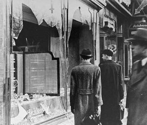 A harrowing photograph showing a Jewish business with a broken window and debris scattered around. Three male onlookers are observing the damaged building, highlighting the aftermath of Kristallnacht and the impact on the local community.