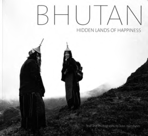 Book cover of "Bhutan Hidden Lands of Hapiness." Book cover has a photograph of two women on a mountain, smiling and wearing a traditional robe and a traditional bamboo hat. 