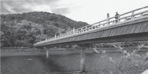  Scenic photograph of Uji River, featuring a bridge with a man standing on it. In the background, a picturesque mountainscape adds to the beauty of the scene.