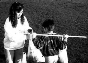 a boy holds grocery bags on each side of a stick he carries on his shoulders as a woman helps him adjust them