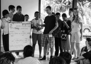 Cambodian and American students sharing knowledge and culture. Students hold a poster at the front of the room and present to other sitting students. 