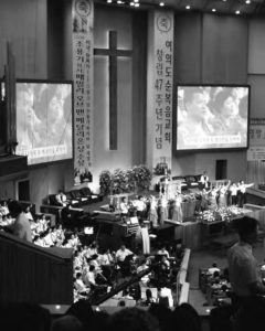 a photograph of the inside of a church. pews full of people can be seen, with large screens and large pillars full of Korean text can be seen on each side of a large cross on the wall.