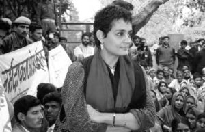 a woman stands with her arms crossed. Behind her are many people, including one holding a banner.