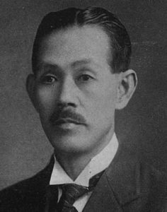 Portrait of Yoshino Sakuzo's face. He is a middle-aged man with thick black hair and a short mustache and is wearing a Western business suit. He is looking away from the camera and into the future symbolizing his role as a visionary and progressive. 