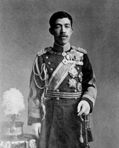 Photograph of Emperor Taisho. He is a middle aged man with thick black hair and a short mustache. He is wearing full military regalia with sash and badges. He holds a feathered military cap in his right hand. 