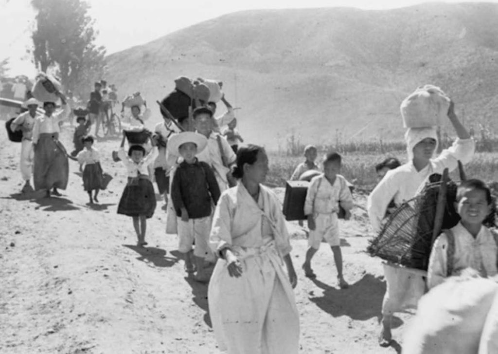  stream of Korean refugees (whether from North or South is unknown) trudges along a dusty road away from the war.The refugees, mainly women and children, are carrying their possessions on their backs or in bundles on their heads.