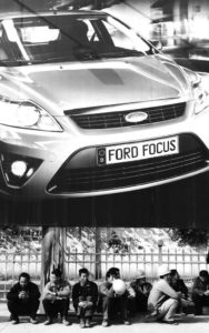 a group of people sitting on the curb in front of a large ford advertisement