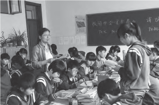 Wang Ping, an Asian woman, standing in front of a student who has risen to answer a question in class. Other students can be seen focused on their open textbooks, actively engaged in learning and participating in the classroom discussion.
