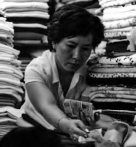 a woman passes money to another person. she stands among stacks of folded garments