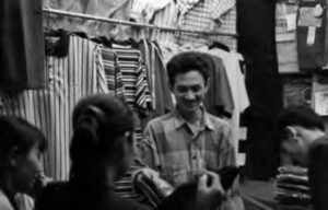 a man smiles and holds folded garments while speaking with some women