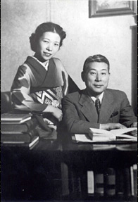 ugihara and his wife Yukiko sitting at a desk, both wearing warm smiles. Yukiko is dressed in a traditional kimono, while Sugihara is wearing a Western-style business suit. Sugihara is holding a book open, suggesting his engagement in work-related activities.