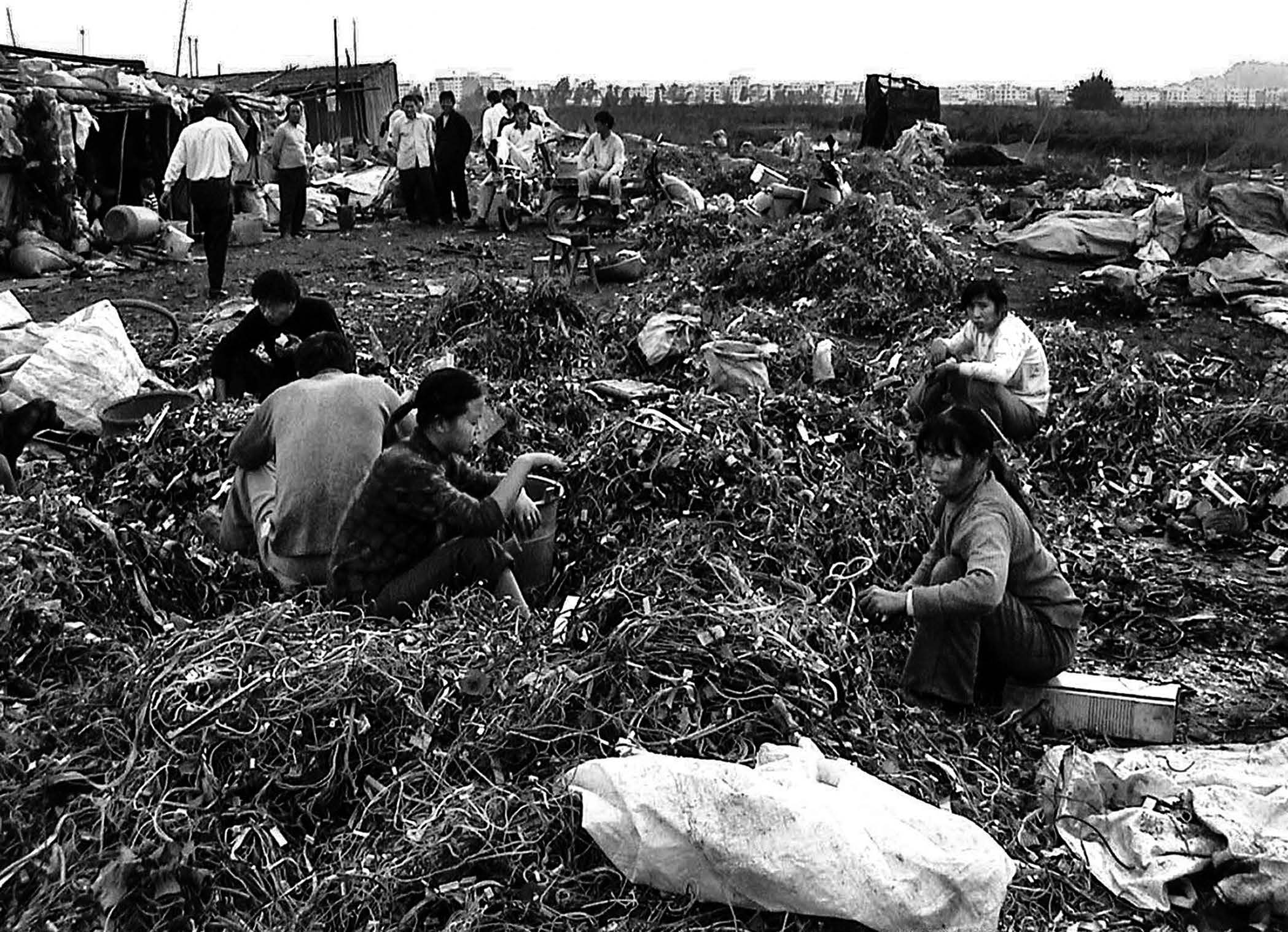 Photograph of factory workers sifting through mounds of wire and electrical waste outdoors. 