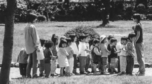 A picture of young children standing in a row in a park while two adults watch them. 