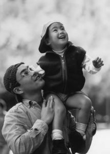 A man holds a young child on his shoulder in a park. They both appear to be staring at something in the sky and smiling. 