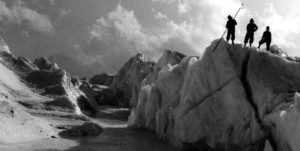 Three sherpa assistants stand on top of a glacier studying it. 