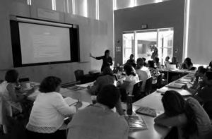 photo of a person teaching at the front board while others in tables listen