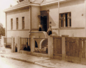 A small building with two individuals standing on the porch. The individuals blend into the background, making it difficult to distinguish specific details about their appearance.