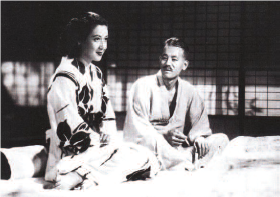 Black and white photograph of Hara Setsuko as Noriko and Ryū Chishūas Shūkichi while on set for the hit film "Late Spring." They are sitting in a tatami room. The woman is smiling and staring at something in the distance, she wears a beautiful white kimono adorned with a flower pattern. The man stares at the woman, he is wearing a plain kimono.