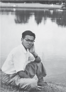 Photograph of 20 year old Hirano sitting by a pond. He is covering his right arm that was covered in Keloid scars following the Hiroshima bombing. He stares intently at the photographer. 