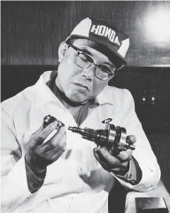 Photograph of middle aged Honda holding a auto mechanic tool and staring intently at it. He wears a laboratory coat, glasses, and a ball cap with the Honda logo. 