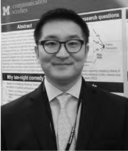 Photograph of a young Hoon Lee wearing a suit and smiling at the camera. He is standing in front of his presentation poster at the University of Michigan. 