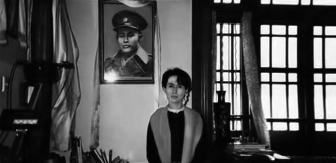 Photo of Aung San Suu Kyi standing in front of a photo of her father, General Aung San, while under house arrest. 