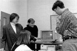 four people crowd around an old computer, pointing at the screen. 
