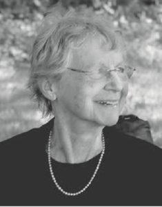 Face photograph of an elderly Jean Elliott Johnson. She has short blonde hair and is wearing glasses and a black shirt with a simple necklace. She is smiling radiantly.