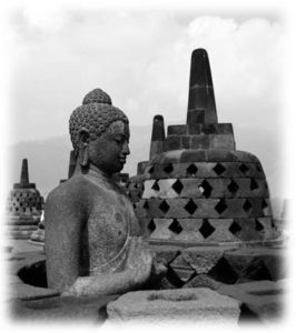 Photograph of a large carved figure of the vairocana buddha and shrines situated near the Merapi volcanoFog clouds the background of the image.