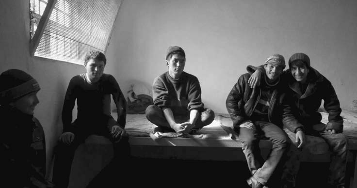 Photograph of migrant workers in Kazakhstan. They are all in a small dorm room and are sitting on a large wooden bed.