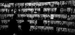 In a museum gallery, two dark figures stand in front of a vast wall covered with hundreds of photographs. The photographs depict individuals who have succumbed to Minamata disease, a condition known to be caused by pollution. The somber scene reflects the significant impact of the disease on the affected community.