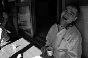 A middle-aged man sits at a table while sipping coffee. He looks up at the camera and smiles.