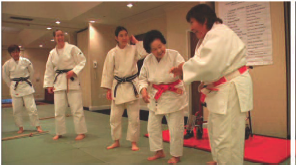 A screen capture from the documentary 'Mrs. Judo: Be Strong, Be Beautiful, Be Gentle.' The image shows five women dressed in Judo outfits practicing in a dojo. The women are engaged in conversation and laughter, displaying camaraderie and enjoyment during their training session.