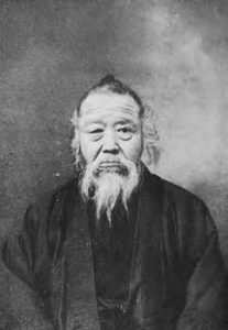 A photograph of Tanaka Shozo captured from the head to the torso. He is an elderly man wearing a modest kimono. He has a long, wry beard. 