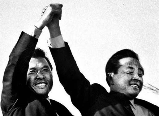 , NDP lawmaker Kim Young-sam (right) holds Kim Dae-jung’s hand high in the air during the campaign tour for the seventh presidential election in Busan. Both men are smiling. 