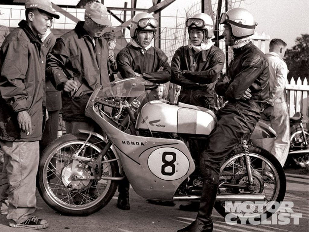 A photograph showing a motorcycle racing team consisting of nine individuals. In the picture, five team members, all middle-aged, are visible. They stand behind a motorcycle, engaged in conversation with a man who is riding a Honda Motorcycle labeled "#8." All individuals in the photograph wear shiny helmets with goggles