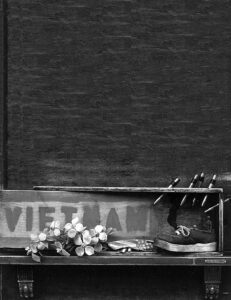 a makeshift memorial: a wooden plank holds an old shoe, a battered american flag, and a bundle of flowers. the wood has "vietnam" painted onto it, and several bullet casings are jammed into the wood.