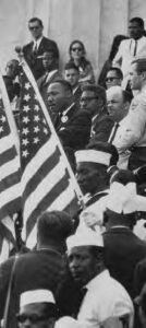 a black man stands at a podium half-obscured by american flags. he is surrounded by many people, both black and white, as he speaks emphatically