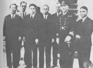 A black and white photograph of seven middle-aged men standing in a row. Six of them are wearing Western-style business suits, while the man standing closest to the camera stands out in a full dress military uniform, adorned with medals and insignia. 