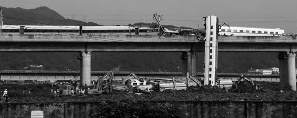 Photograph of a train crash on a bridge. One train car appears to have completely derailed and is hanging off the side of the bridge. 