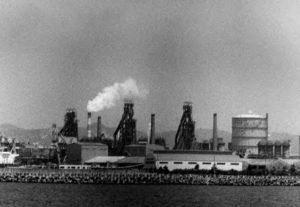 large metal spokes reach above a city, with one spewing smoke