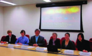 A group of students and faculty sit in a line delivering a powerpoint presentation. 